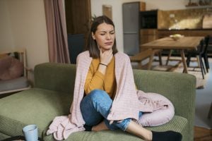 woman suffering from sore throat sitting on sofa. Sick young woman covered with blanket coughing while sitting on the sofa at home.